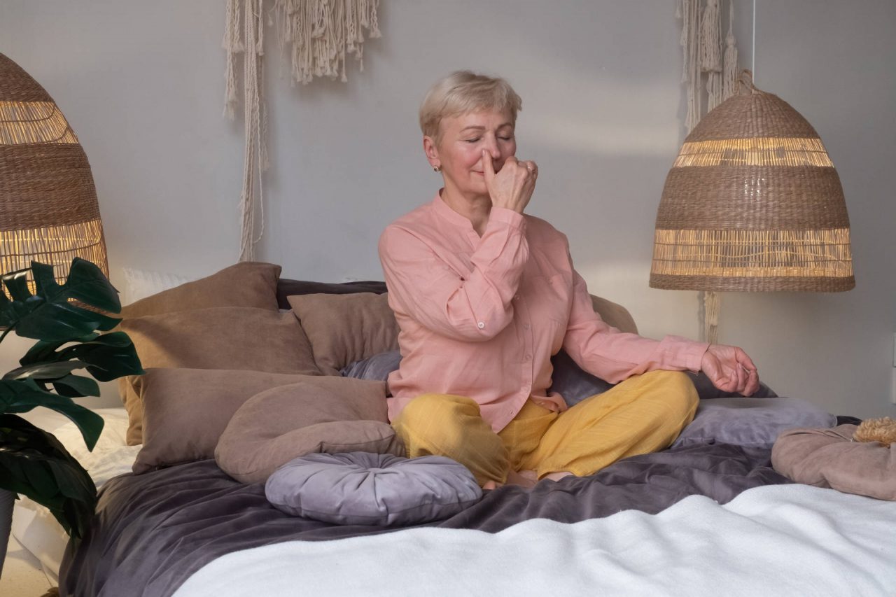 a senior woman practising yoga at home, sitting on a bed breathing with pranayama